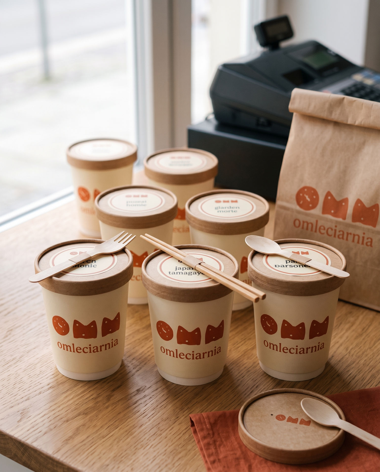 six omelettes packed in branded takeaway cups with wooden lids, a wooden fork, chopsticks and a spoon banded to the front three lids, a kraft paper bag beside them